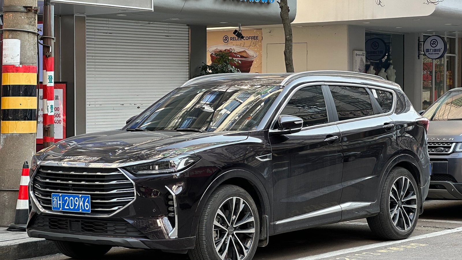 JETOUR X90 PLUS de color oscuro estacionado en una calle comercial de China, visto de frente y de lado